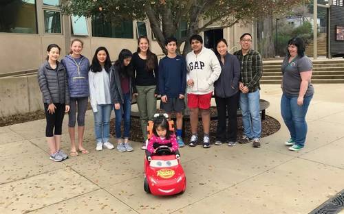 Toddler in a modified ride-on car surrounded by middle school student engineers